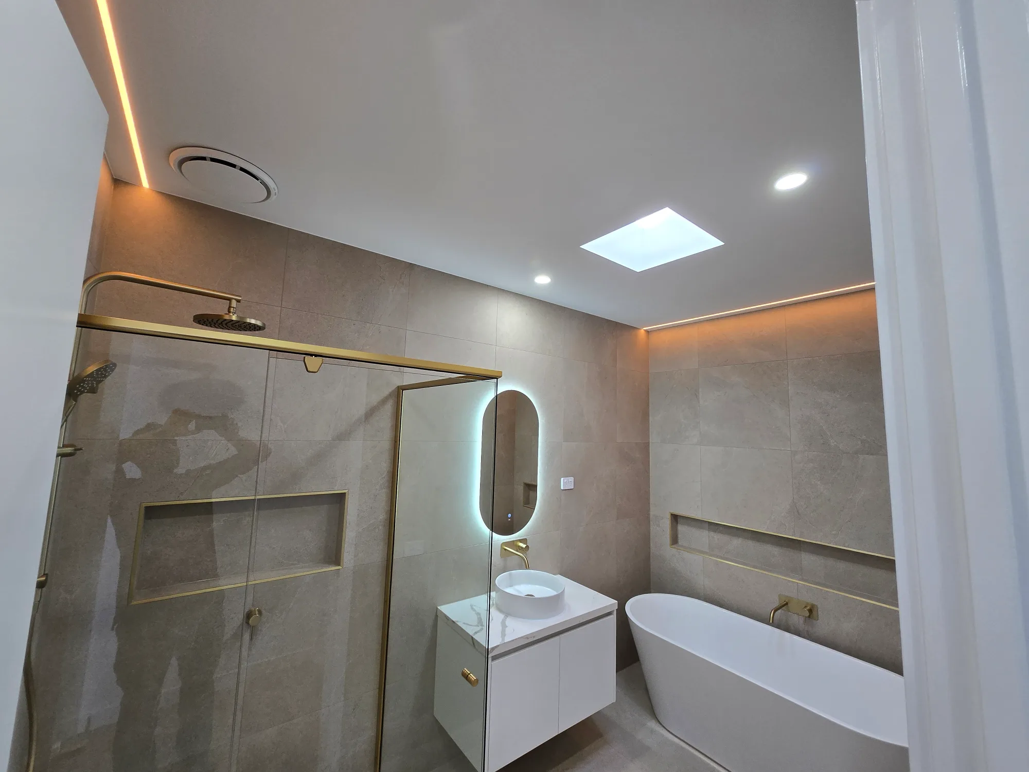Luxury bathroom in a new Melbourne home featuring a skylight, freestanding bathtub, frameless shower screen, custom vanity, large-format tiles, and brass fixtures.